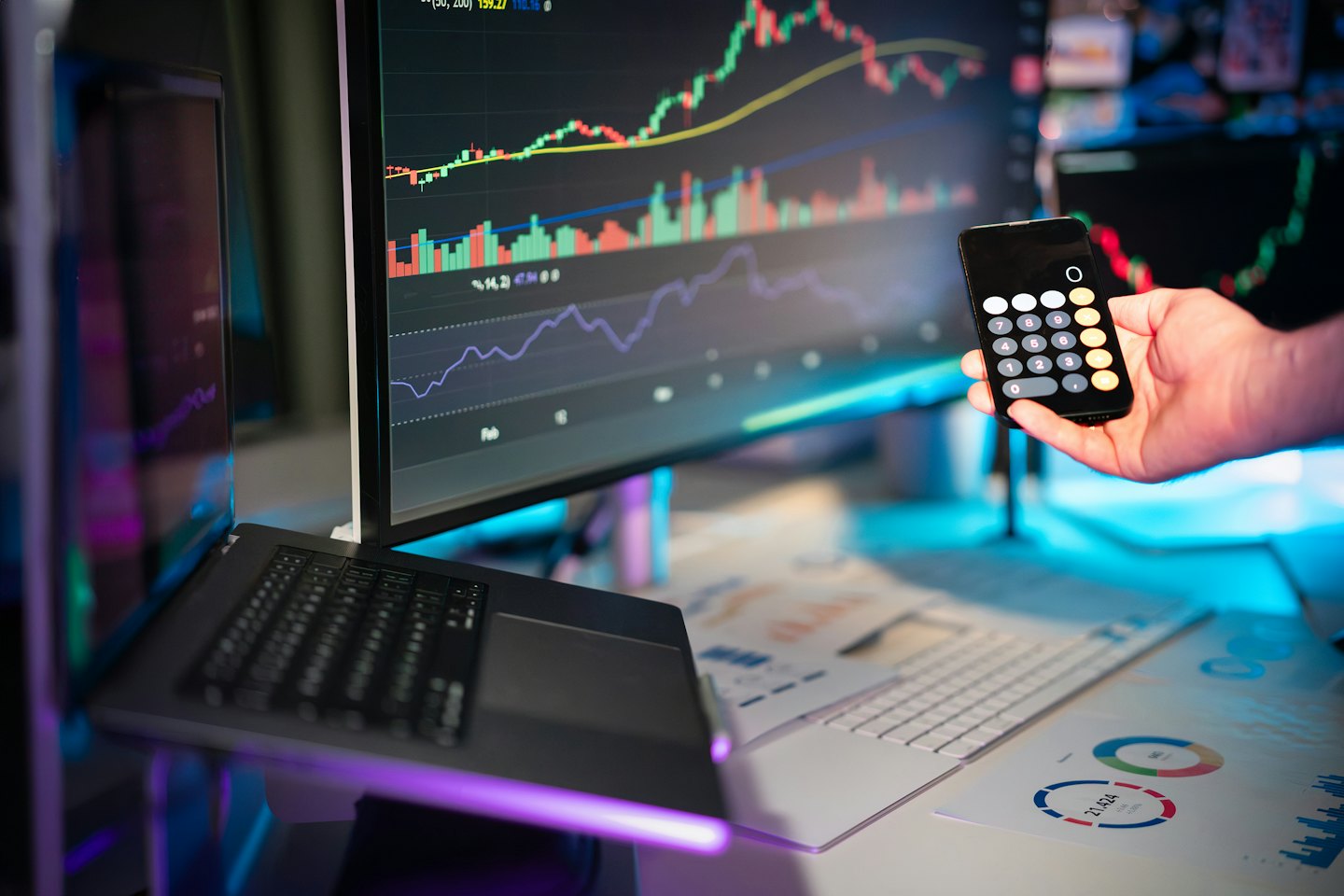 Servizi Mano che tiene un telefono con una calcolatrice vicino a dati finanziari su monitor.